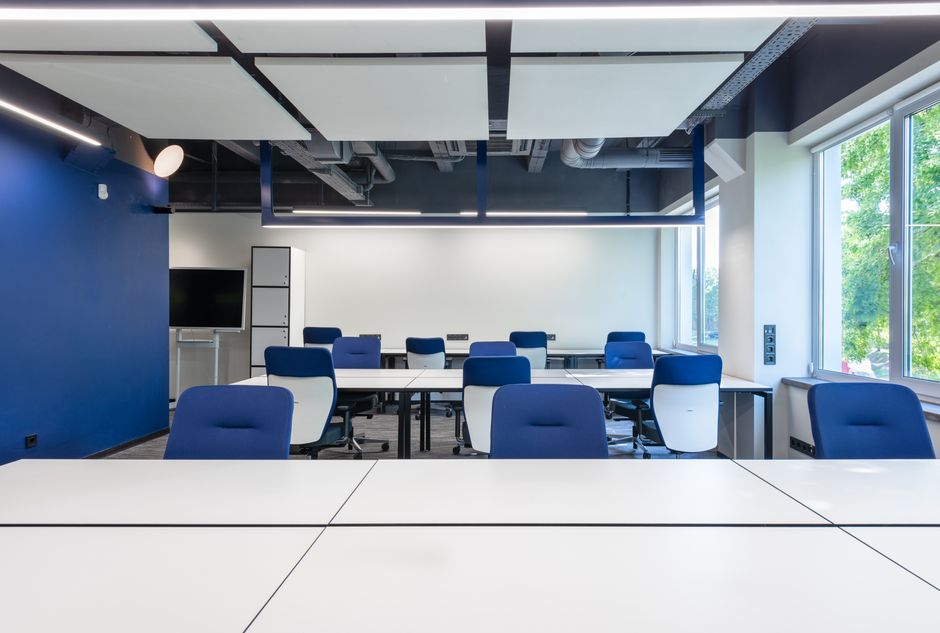 Various white desks with blue chairs placed in modern spacious workspace with big window and monitor in contemporary business center.